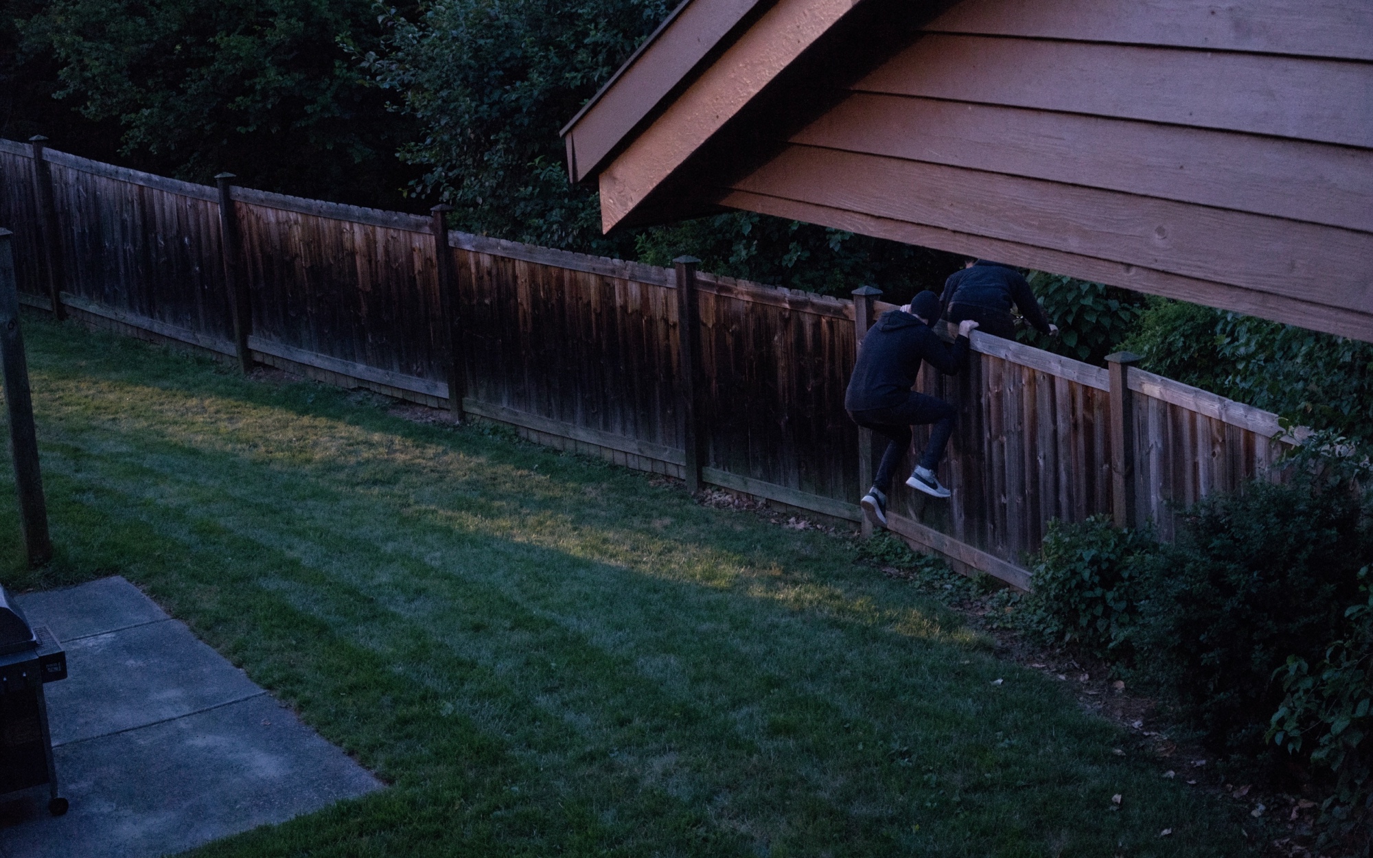 Backyard security-camera view at dusk looking down at the fence line: figures climbing over the back fence into the yard